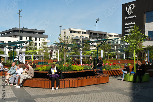 People relax in the modern outdoor area of Posnania shopping center in Poznań, Poland, surrounded by flowers, benches, and urban architecture.