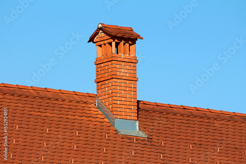 Fotografie Tall red brick chimney with decorative cap rises from a red clay tile roof, with