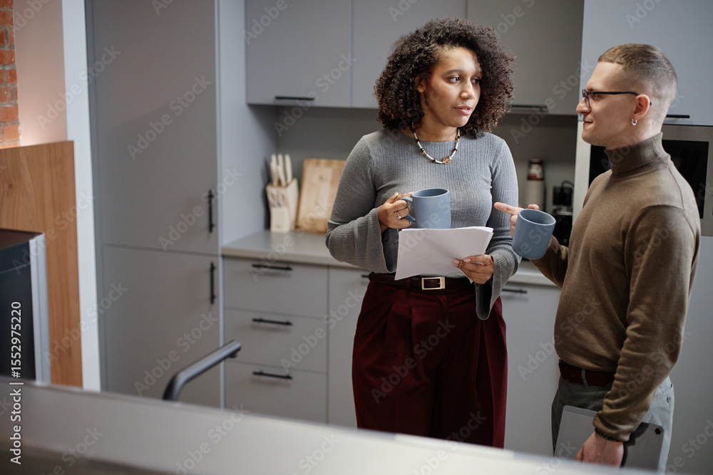 Obraz premium Caucasian young adult man and Black woman standing in modern office kitchen holding mugs and documents, engaging in conversation, woman looking away from camera