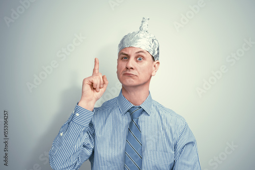Suspicious man in striped shirt and tie wearing a DIY tin foil hat, raising finger with intense expression. Conspiracy theorist concept on light grey background.