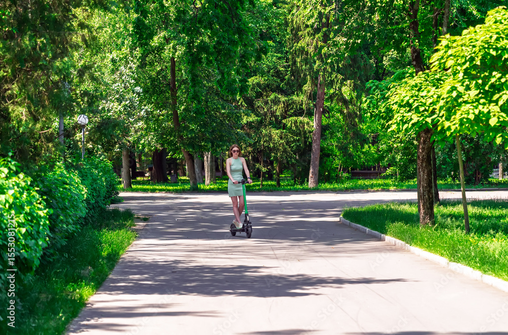 Fototapeta premium Modern urban transport: A stylish girl in a dress enjoying a ride on an e-scooter in nature.Summer leisure and active lifestyle. A woman on a kick scooter in a public city park.Eco-friendly commute