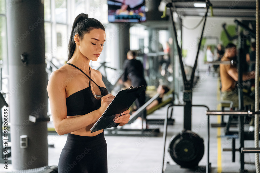 Fototapeta premium With notepad. Female trainer is standing in the gym