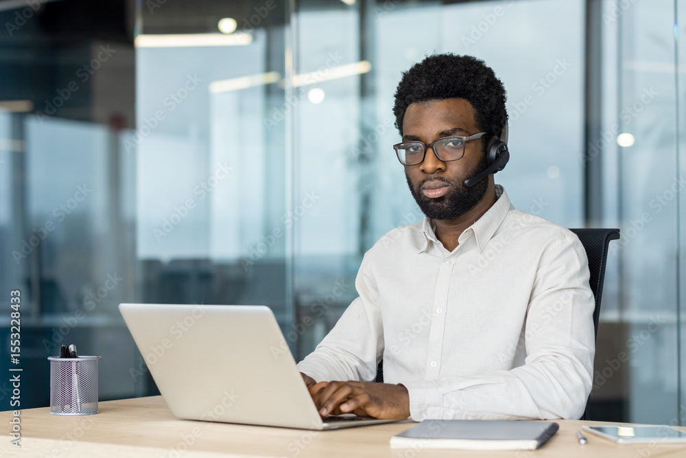Fototapeta premium An African-American man wearing a headset and glasses works on a laptop at a desk in a modern office setting.