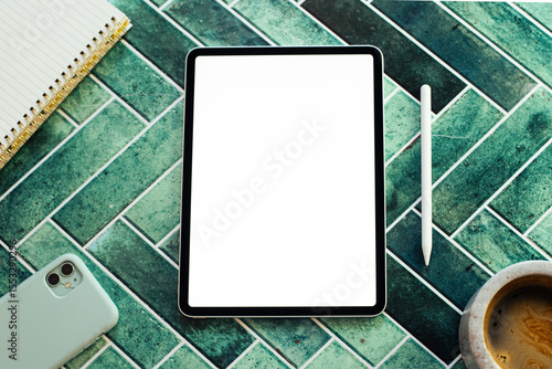 Top view of a digital creative workspace featuring a blank screen tablet, stylus, notebook, smartphone, and a cup of coffee on a stylish green tile surface. Ideal for concepts like remote work