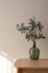 Green vase with olive plant branch on wooden table top, on neutral beige plaster wall background with natural sunlight shadow, aesthetic minimal home interior decor