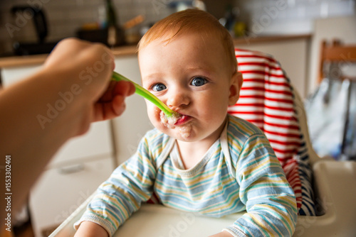 little cute boy 8 months old with blond hair and blue eyes eats porridge with a spoon in a high chair