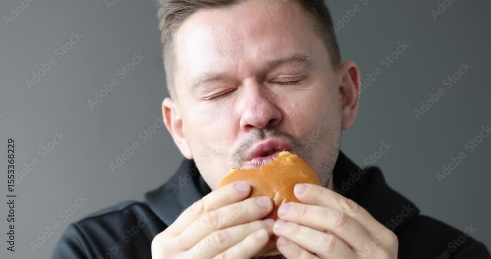 Man with closed eyes savors delicious cheeseburger holding food close to face. Guy gets completely immersed in taste and juicy fast food aroma Man with closed eyes savors delicious cheeseburger holding food close to face. Guy gets completely immersed in taste and juicy fast food aroma