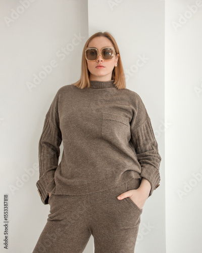 young stylish plus size blonde girl in brown casual costume is standing fashion and looking straight in sunglasses on the white art wall background. teen fashion. fashion concept, free space