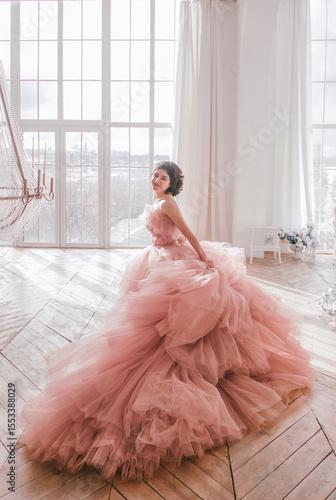 young girl with short black hair in pink princess dress with train is standing from the back and smiling cute near huge panoramic white window background at sunny day. fashion concept, free space