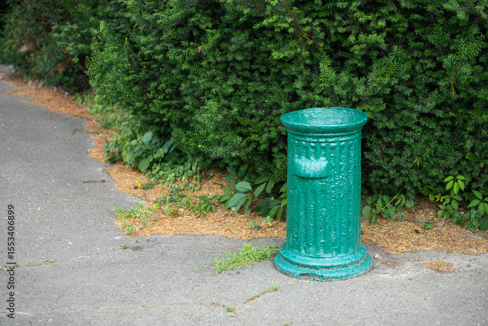 Fototapeta premium A weathered green trash can stands beside a quiet pathway in a park, surrounded by vibrant greenery and scattered mulch. The atmosphere is calm and inviting for visitors