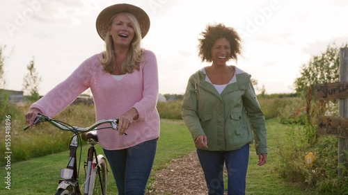 Two Mature Female Friends Walking Along Path With Bike Through Yurt Campsite
