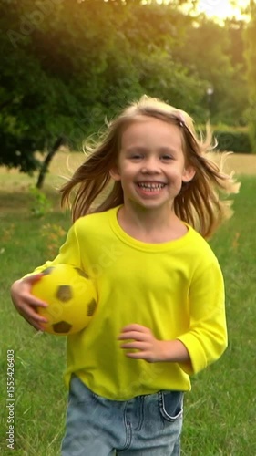 Camera follows on running happy child with soccer ball on green lawn in summer park at sunset. Little girl plays and laughs outdoors. Football and sport. Vertical shot