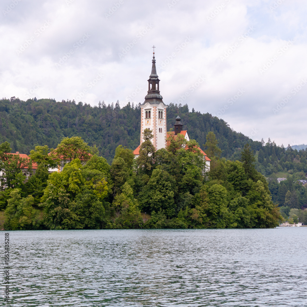 A serene view of the iconic Pilgrimage Church of the Assumption of Mary situated on Bled Island, rising above lush green trees in the middle of Lake Bled A serene view of the iconic Pilgrimage Church of the Assumption of Mary situated on Bled Island, rising above lush green trees in the middle of Lake Bled