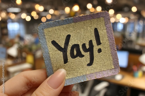 A hand holding a yellow sticky note with the word “Yay!” written in purple marker, in a colorful modern office background, ideal for concepts of celebration, achievement, or motivation.