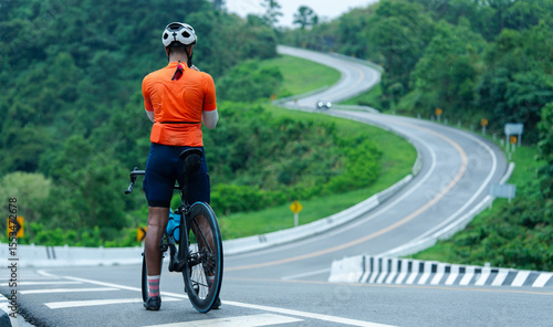 A cyclist in orange gear stands beside a road bike, overlooking a winding road surrounded by lush green foliage and trees.