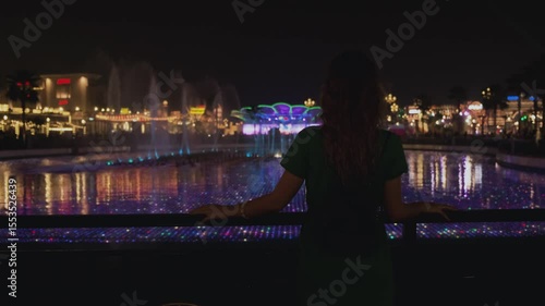 Delightful bewitching sight singing musical multicolored fountains Arab resort city Dubai. Young woman tourist turned away, enjoy beautiful panoramic view, colorful illumination night city.uae 4k 2020