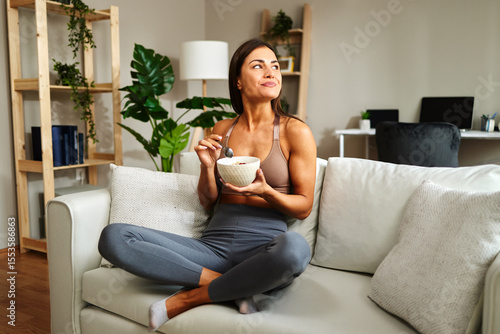 Young sportswoman eating healthy breakfast at home while sitting on sofa