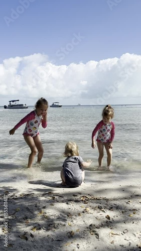 Three kids playing together in shallow sea water on sunny day, joyful siblings enjoying beach vacation, splashing waves, sand, boats in background, carefree childhood fun and laughter at seaside