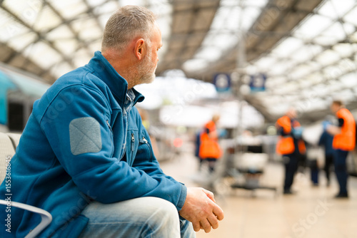 Man waiting in a busy train station during daytime, observing staff in bright uniforms assisting passengers nearby