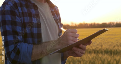 Close-up of male farmer holding pen and clipboard, writing notes during crop inspection in wheat field at sunset. Concept of agricultural documentation, crop control, and farm work.