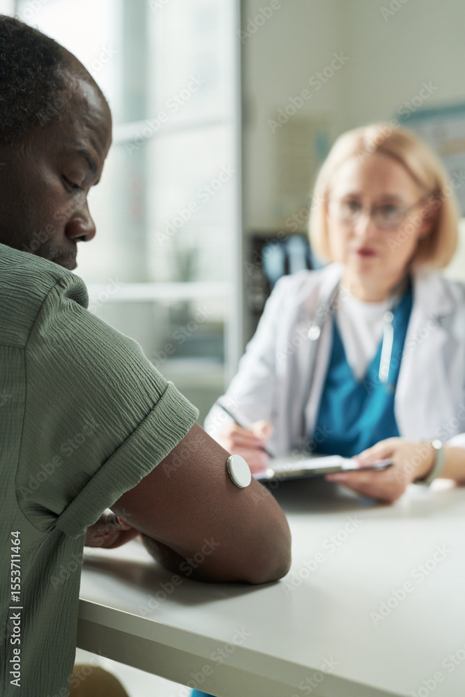 Obraz premium Middle aged Black man wearing glucose monitor on arm consulting with Caucasian female doctor in medical office, doctor holding clipboard and discussing diabetes management