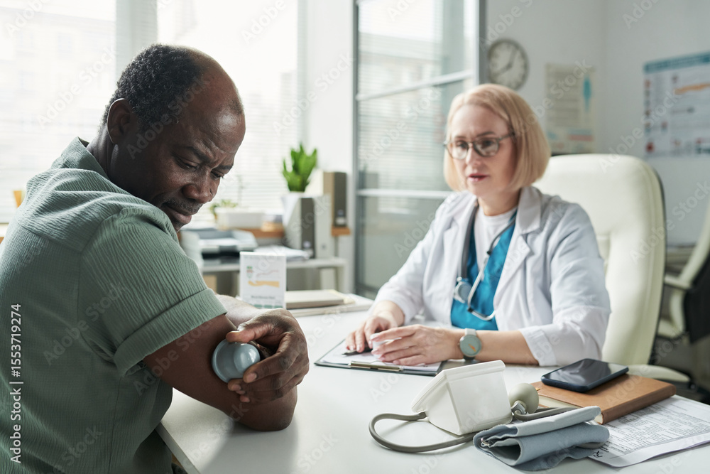 Obraz premium Middle aged Black man checking blood glucose with glucometer while sitting across from middle aged Caucasian female doctor in medical office during diabetes consultation