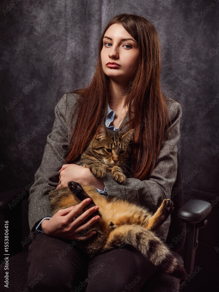 Obraz premium Classic beauty portrait of a young girl in grey business suit holding cat on grey painted background. Fashion and beauty. Model has long hair and calm confident face. Feminine soft pose. Relaxed mood