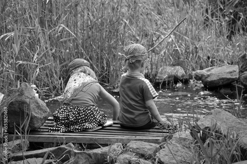 Horizontal black and white photo of two children sitting on a wooden bridge near a small stream, surrounded by tall grass and rocks. Nostalgic and peaceful moment of childhood in nature.