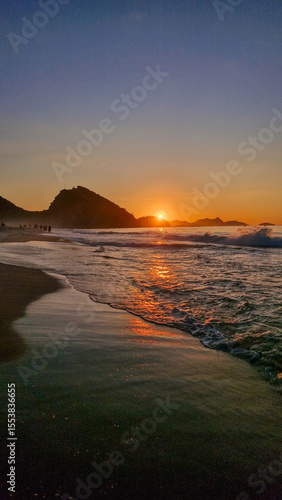 Lindo nascer do sol em uma praia com montanhas ao fundo e um céu com um gradiente de azul para laranja e mar calmo refletindo a luz do sol.