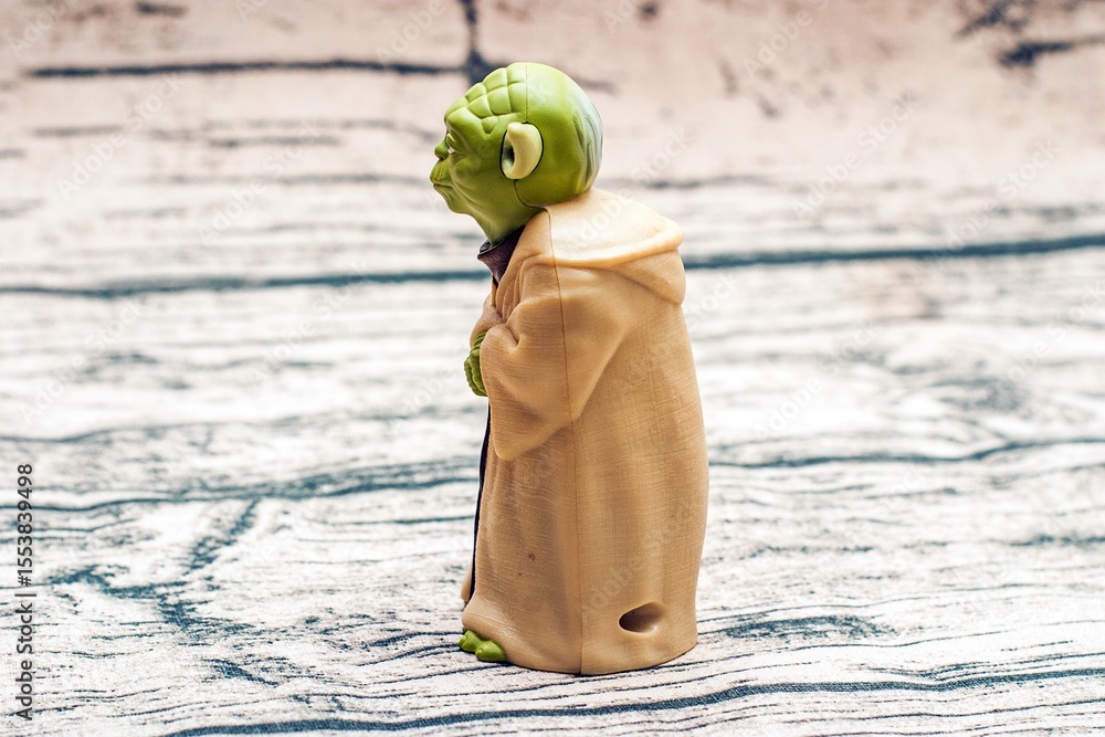 Naklejka premium A small green Yoda figurine dressed in a beige robe stands on a textured surface