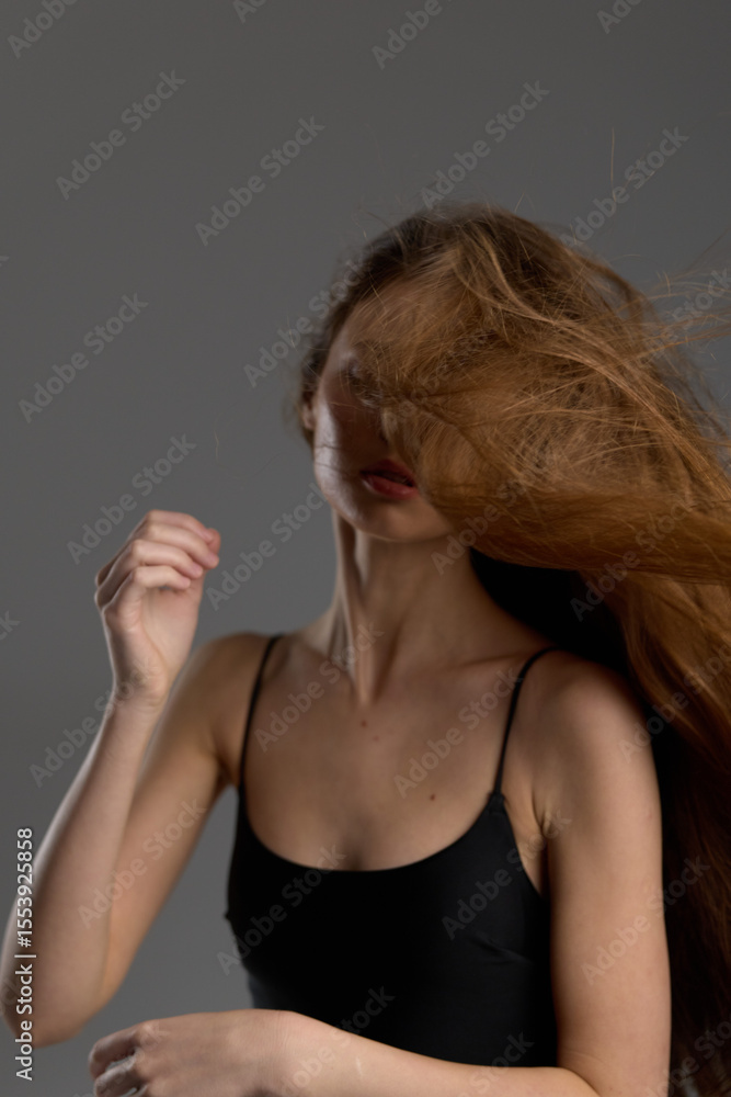 Obraz premium woman with long hair in black tank top moves hair dynamically against gray background, capturing motion and natural beauty in soft lighting.