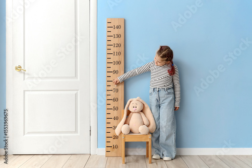 Little girl measuring toy's height on chair near stadiometer at home