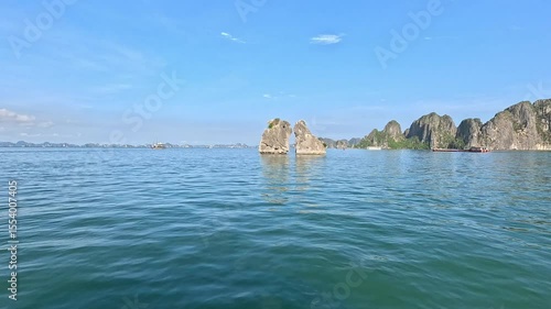 A scenic view of the limestone islets known as the Kissing Rocks, rising from the calm emerald waters of Ha Long Bay, Vietnam, under a clear blue sky.