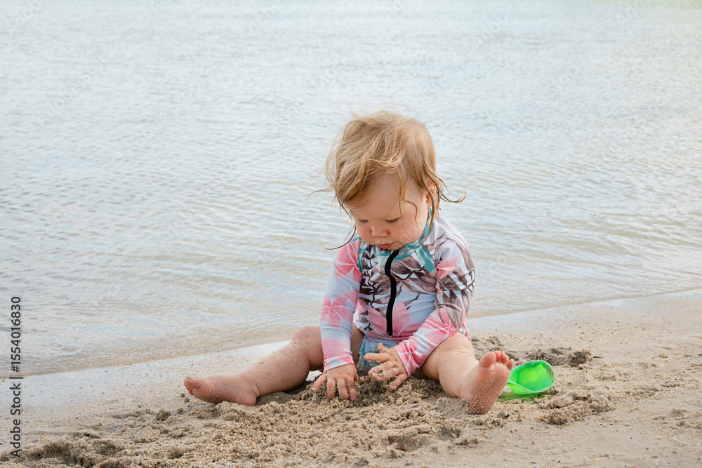 Adorable toddler girl sitting at the edge of the sea on sandy beach, wearing colorful sun protective swimsuit, playing with wet sand, exploring shoreline, carefree childhood summer vacation moment out Adorable toddler girl sitting at the edge of the sea on sandy beach, wearing colorful sun protective swimsuit, playing with wet sand, exploring shoreline, carefree childhood summer vacation moment out