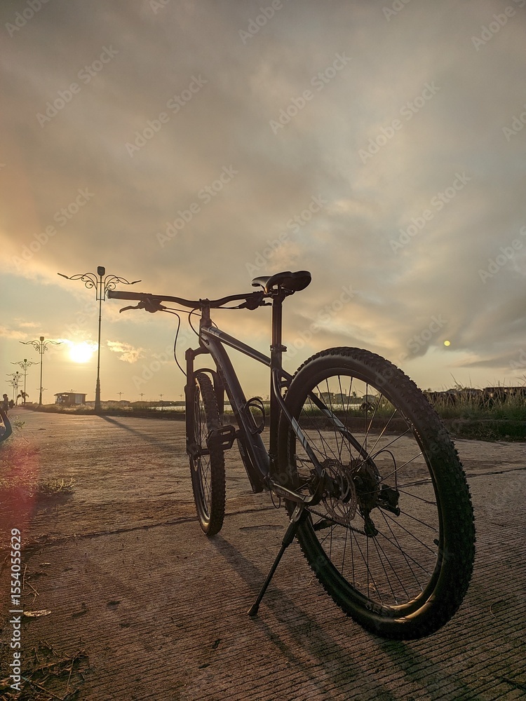 Obraz premium A bicycle sits parked on a concrete path during a golden sunset, casting long shadows around it.