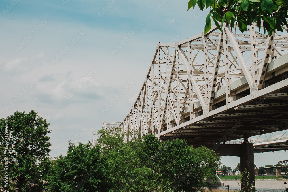 Fototapeta premium Steel truss bridge spanning a river framed by leafy trees under a clear blue sky in a serene outdoor environment