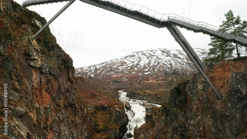 Impressive Voringsfossen footbridge spans over deep gorge with winding river against snowy mountains. Dramatic natural landscape in Norway region