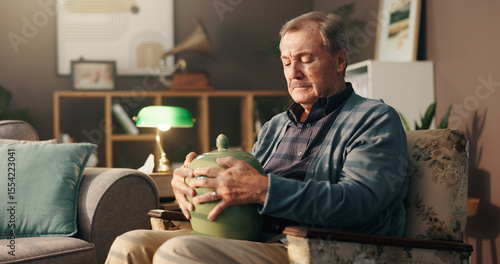 Senior, sad man and home with urn or memory for grief, loss or mourning in thought on chair. Elderly, male person or widower thinking with vase of ashes for nostalgia, missing piece or lonely house