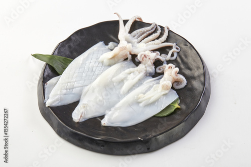 Fresh raw squid arranged on black ceramic plate with green leaves, showcasing its tentacles and body parts, ready for cooking or seafood preparation