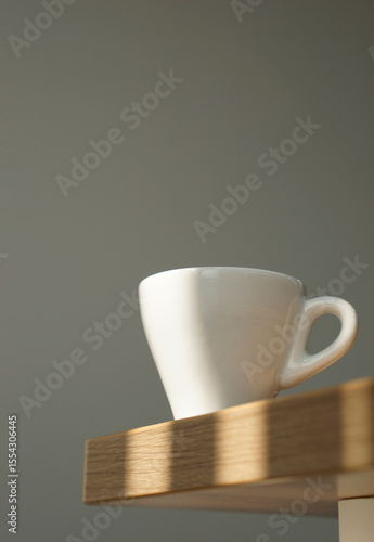 Close up white coffee cup on wooden table under morning window light on clean grey wall background in vertical.