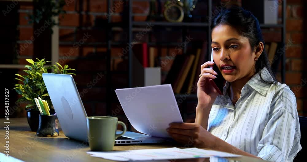 Businesswoman attending phone call with documents at desk in modern cozy office, Indian Asian female holding reports while communicating on mobile during office hours with confident expression Businesswoman attending phone call with documents at desk in modern cozy office, Indian Asian female holding reports while communicating on mobile during office hours with confident expression