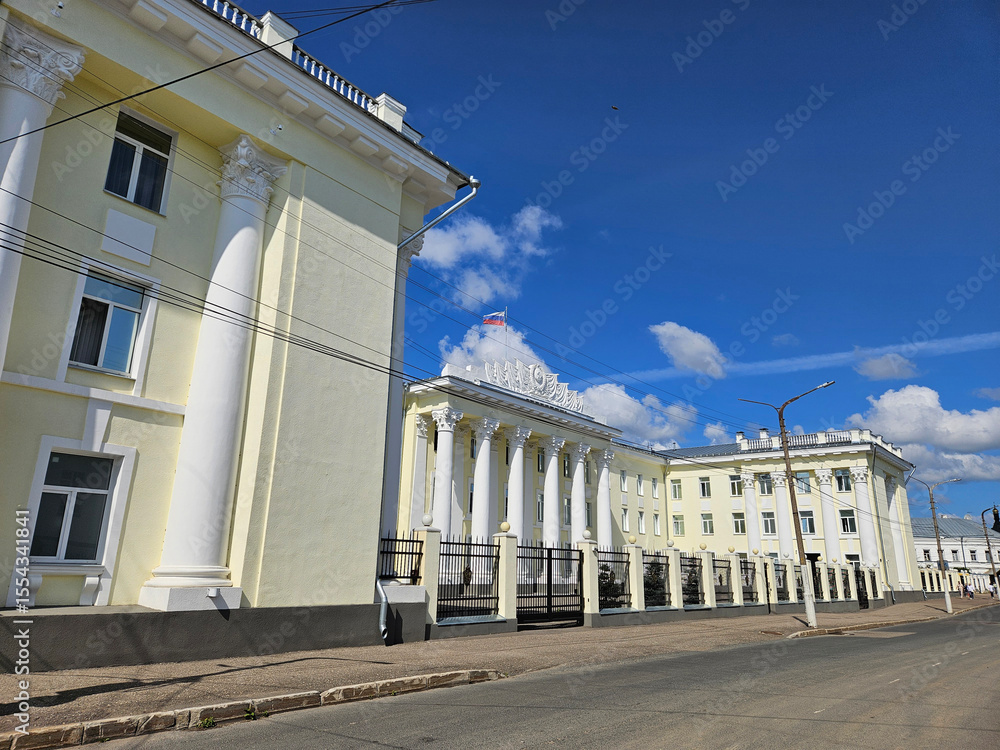 Fototapeta premium Large building Office of the Federal Security Service for the Kostroma Region with a white facade and a red and white flag on top. The building has a lot of columns and is surrounded by a sidewalk