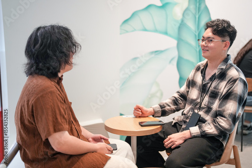Two Chinese men in their early twenties, both wearing glasses, are sitting together in a stylish cafe, casually talking and laughing. It is a warm May afternoon in Huangpu District, Shanghai, China.