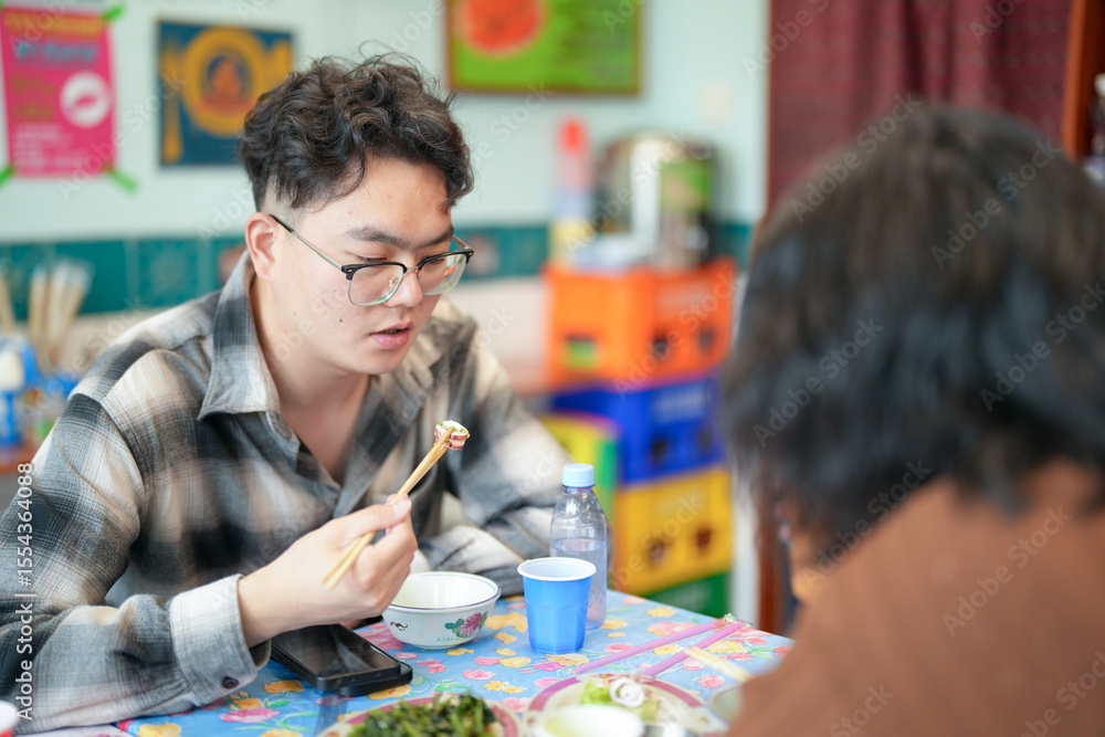Fototapeta premium Two young Chinese men in their early twenties, both wearing glasses, are eating lunch together at a Thai restaurant, enjoying a casual spring afternoon in Huangpu District of Shanghai, China.