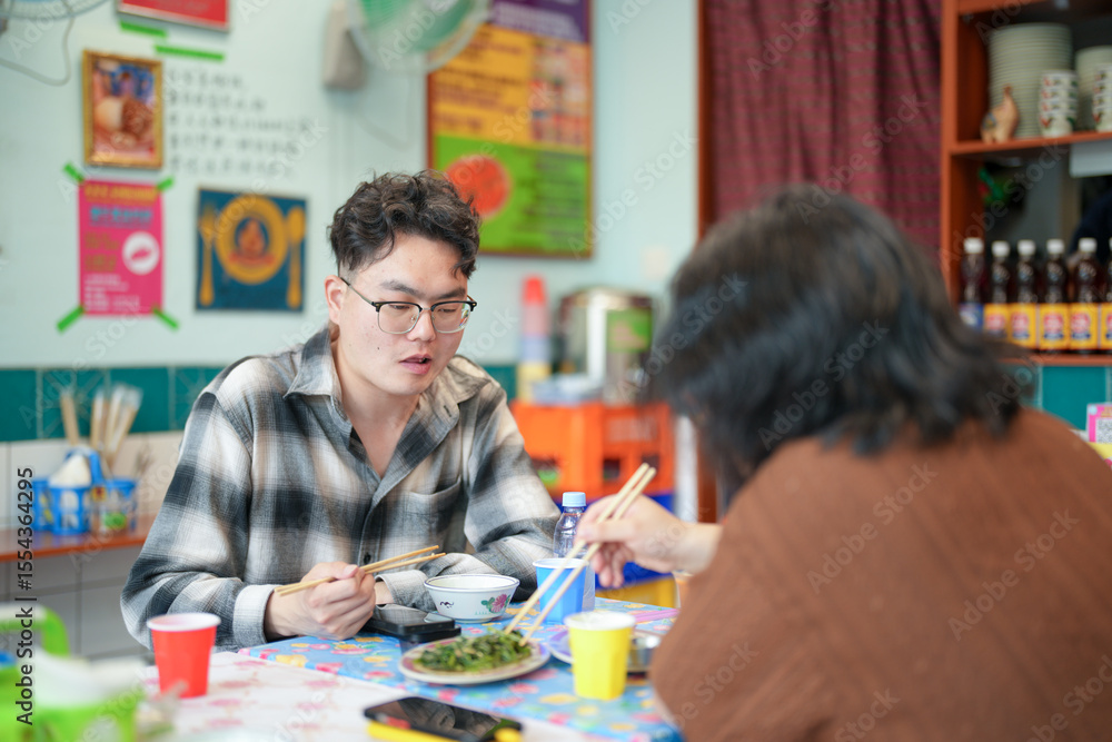 Fototapeta premium Two young Chinese men in their early twenties, both wearing glasses, are eating lunch together at a Thai restaurant, enjoying a casual spring afternoon in Huangpu District of Shanghai, China.