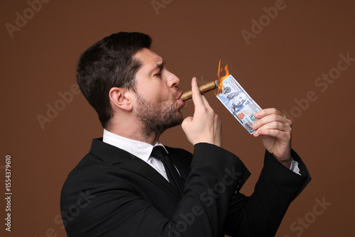 Fototapeta Greedy businessman lighting cigar with dollar banknote on brown background