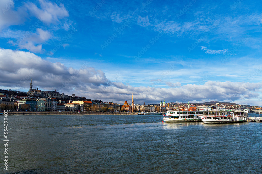 Obraz premium Panoramic view of the Danube River with historic buildings and boats in Budapest, Hungary under a vibrant blue sky.