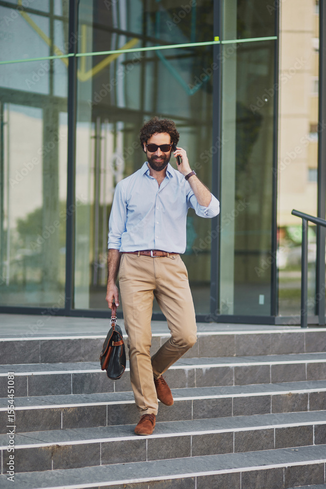 Fototapeta premium A man dressed in a light blue shirt and beige pants is walking down the steps of a modern office building while speaking on his mobile phone. He holds a stylish bag and looks focused.