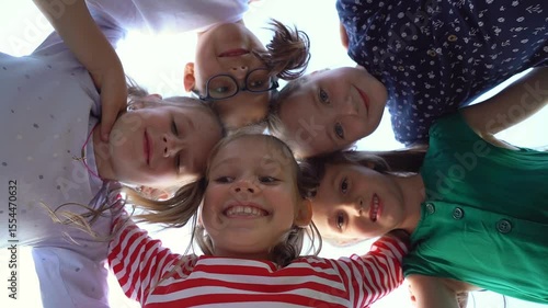 Low angle view of happy little children in circle look at camera embrace and laugh in summer park. Team kids play outdoors. Close up of face girls and boys. Schoolchildren on holiday