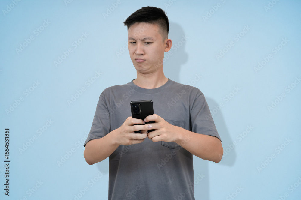 Young Asian Man Looking at Phone with Thoughtful Expression, Pondering Message, Light Blue Background Young Asian Man Looking at Phone with Thoughtful Expression, Pondering Message, Light Blue Background